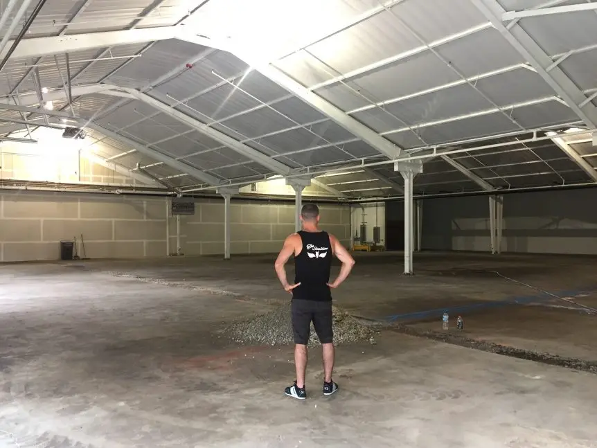 Man in tank top stands in large empty warehouse space with concrete floor, looking at future gym site.
