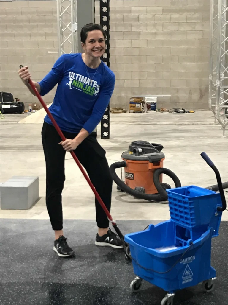 Woman in Ultimate Ninjas shirt mops a gym floor with a blue mop bucket and shop vac in the background.