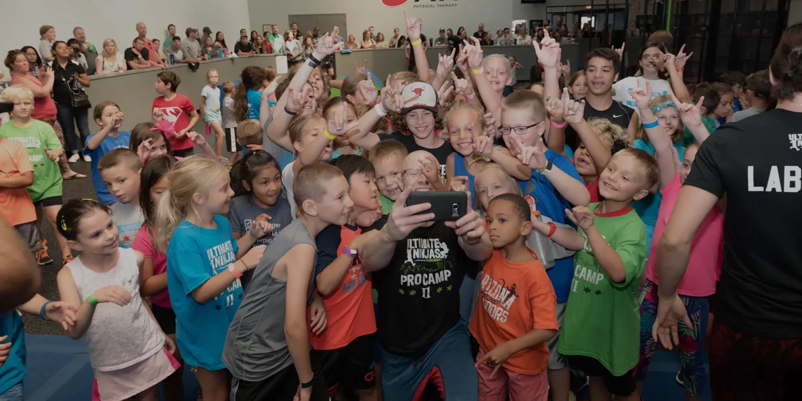 A male Ultimate Ninjas coach takes a selfie with a large group of smiling children, many making hand gestures.