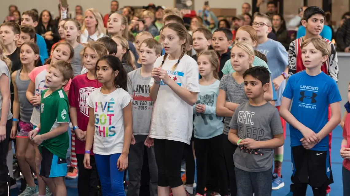A large group of children and adults stand attentively at a kids ninja gym.