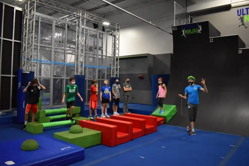 Instructor teaching kids in a ninja gym class, near red Angled Steps and green balance blocks.