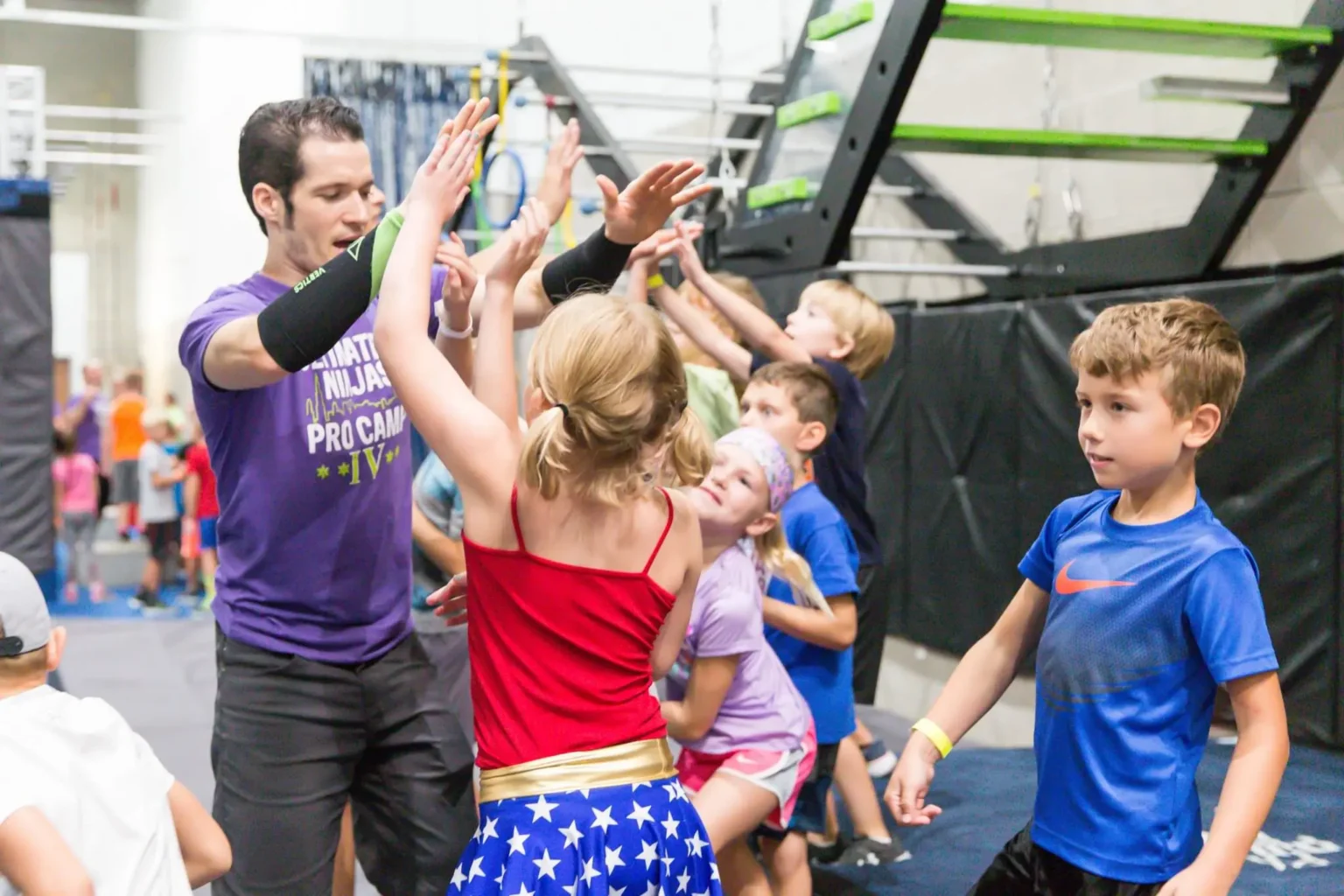 Ultimate Ninjas coach and kids high-fiving, with angled Devil Steps obstacle in the background.