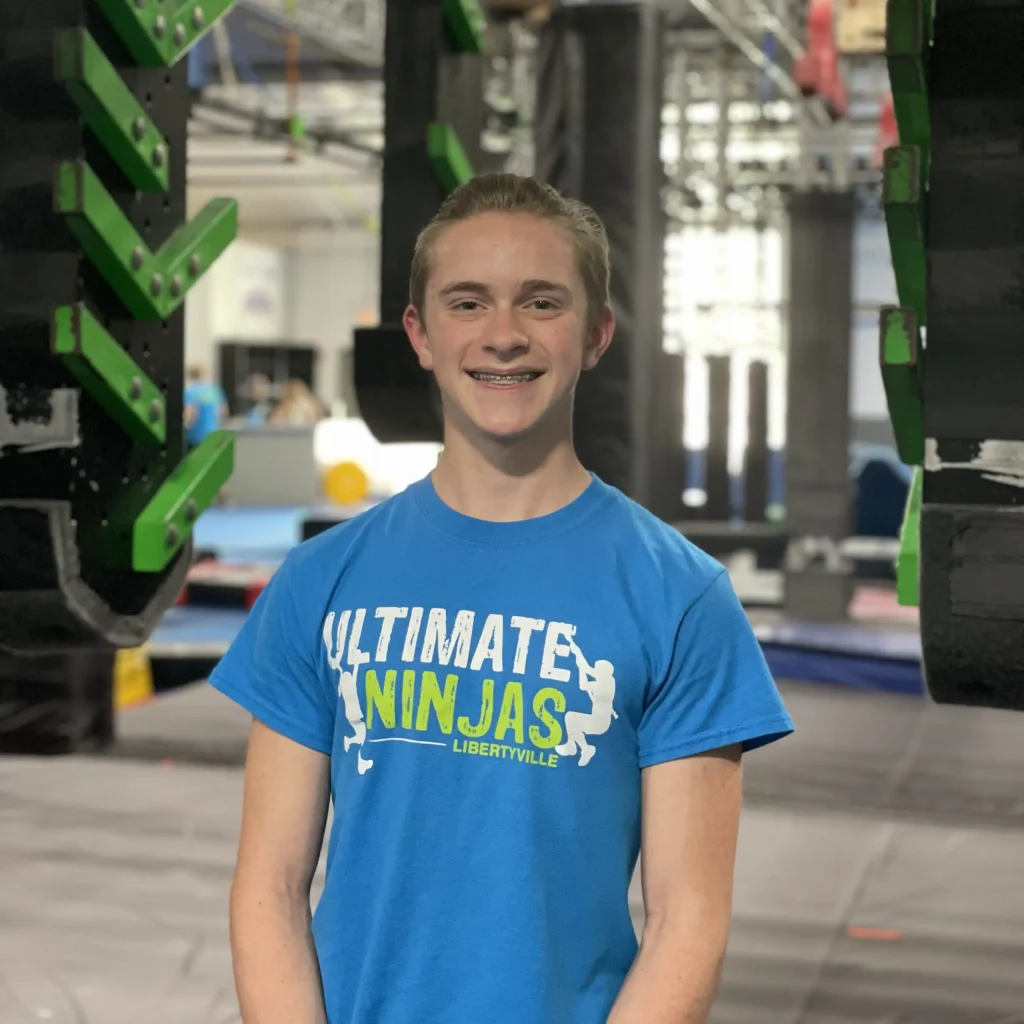 Smiling boy wearing an Ultimate Ninjas t-shirt standing inside a ninja gym.