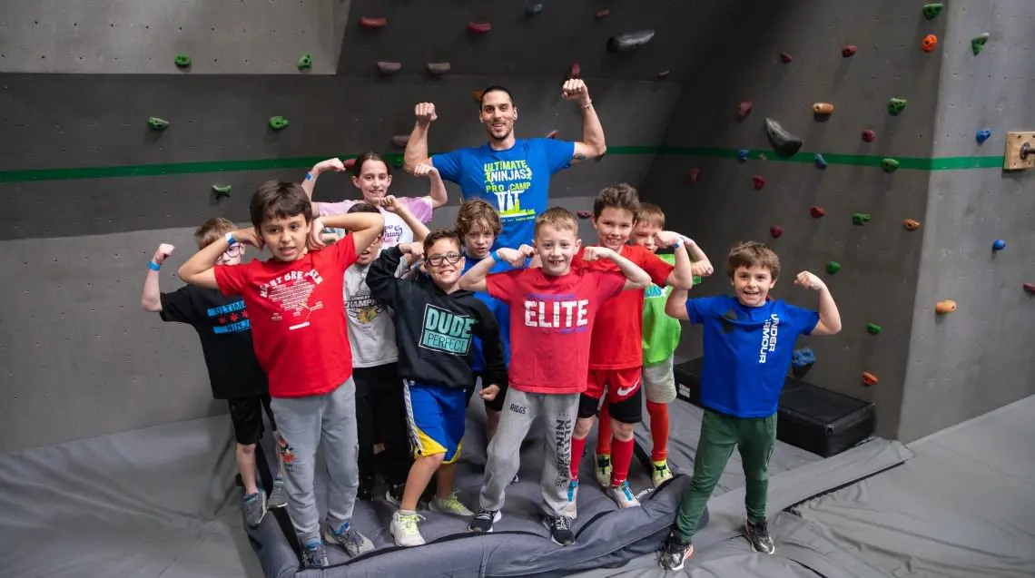 Ultimate Ninjas coach and group of kids flex muscles on soft mats in a ninja gym.