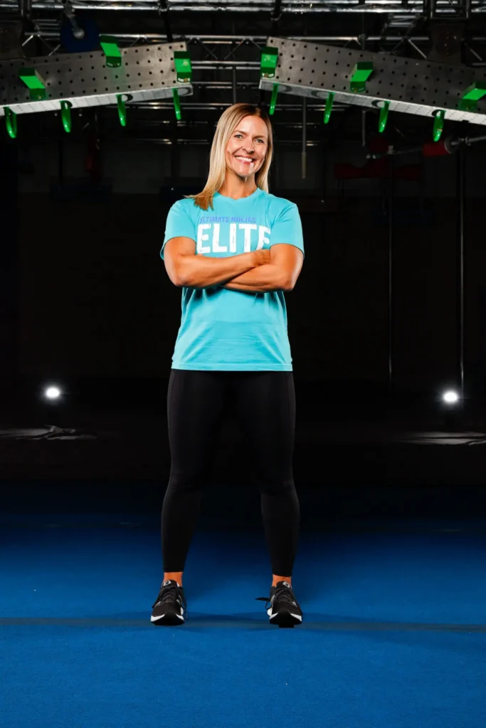 Woman in Ultimate Ninjas Elite shirt, smiling with arms crossed in a gym.
