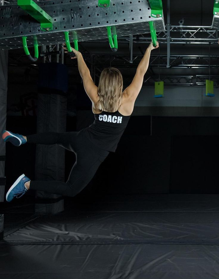 Coach hanging from two green rings, legs extended, demonstrating a grip line obstacle.