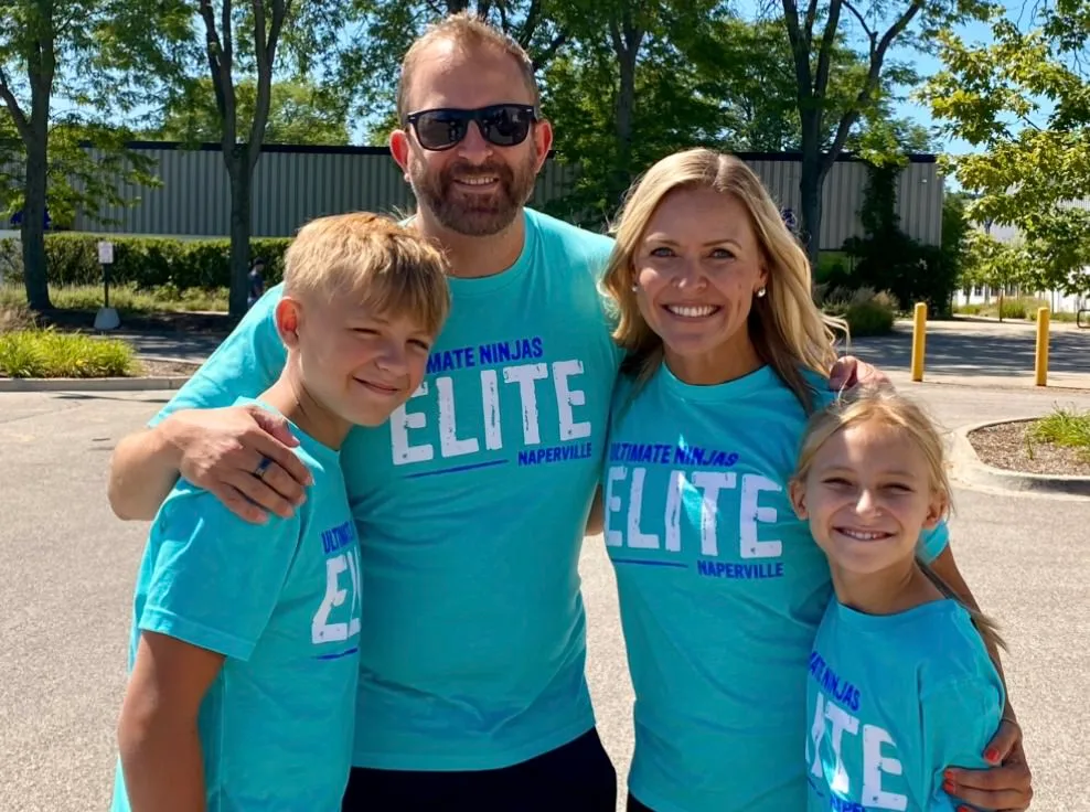 Family of four, two adults and two children, wearing matching Ultimate Ninjas Elite shirts, smiling outdoors.