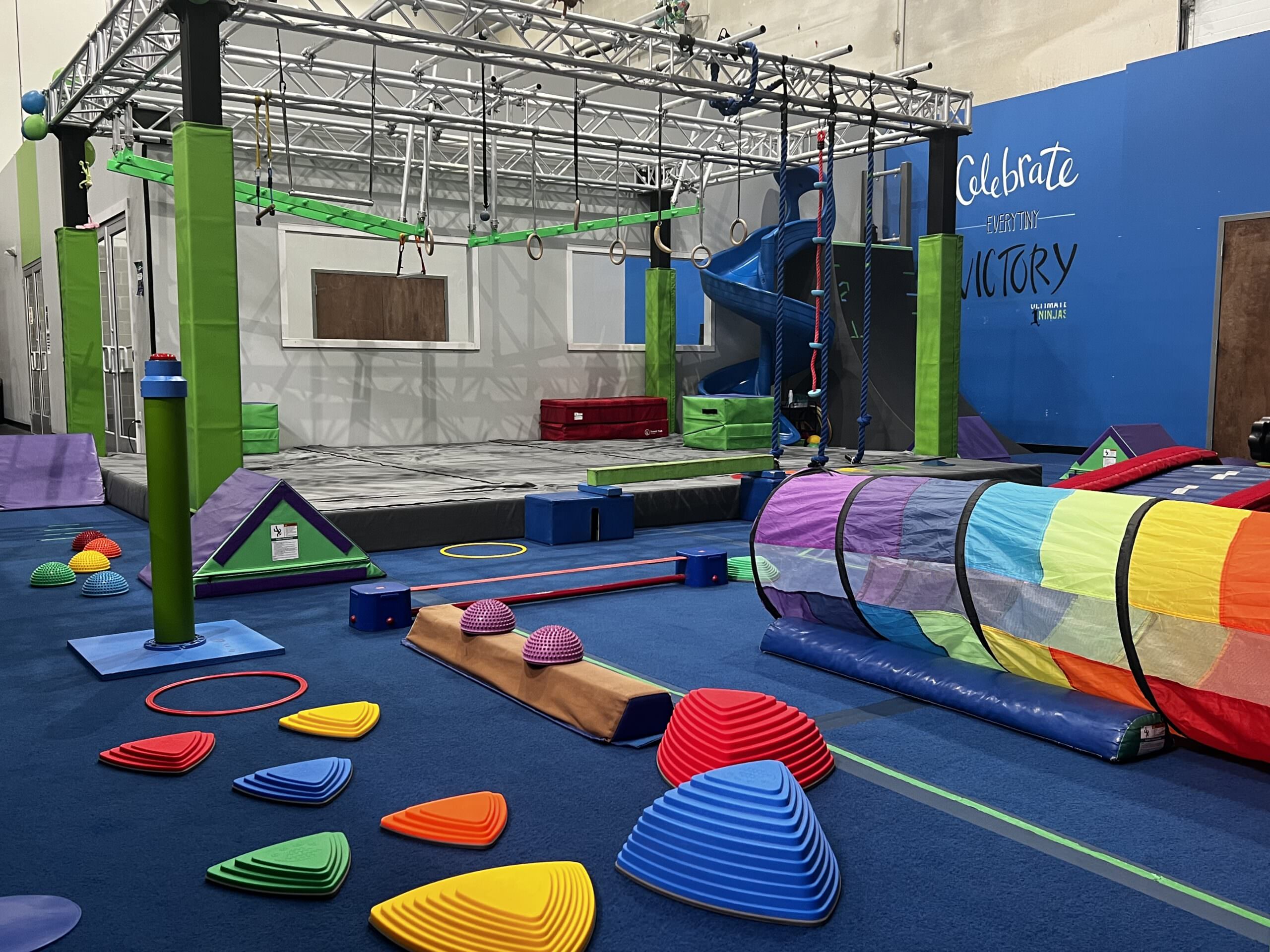 Kids ninja gym featuring an overhead rig with bars & angled steps, colorful balance obstacles, crawl tunnel, and slide.