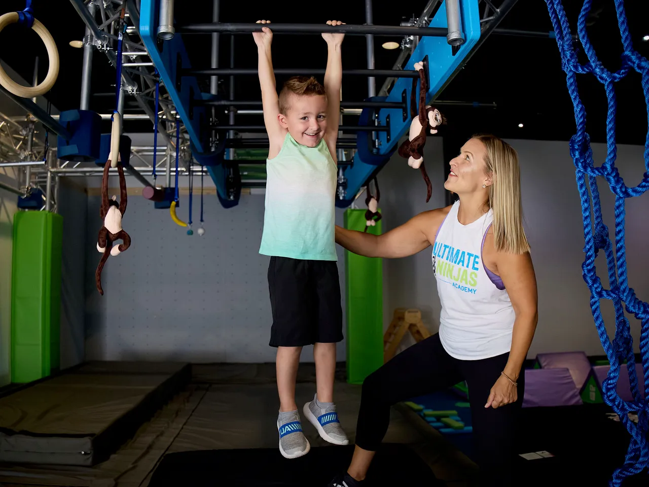 A smiling young boy hangs from a blue monkey bar, while a female Ultimate Ninjas coach assists him.