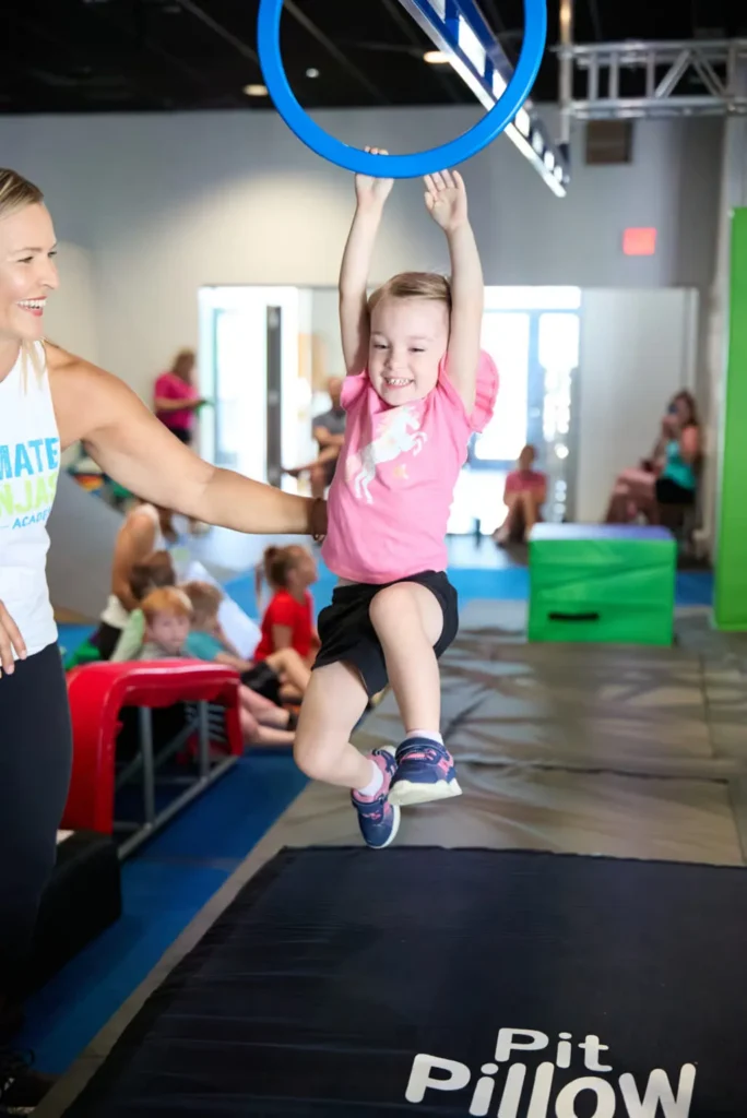 Smiling child swings from a lache lane bar, helped by an instructor, over a trampoline pad.