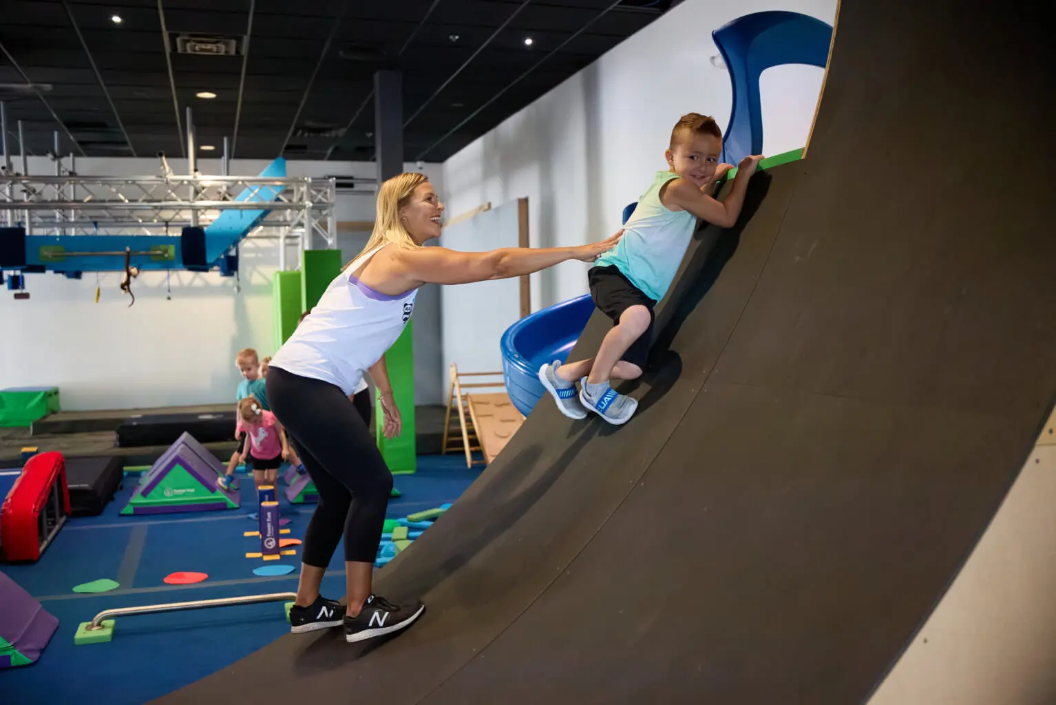 Woman helps a young boy climb a Warped Wall in a ninja gym.