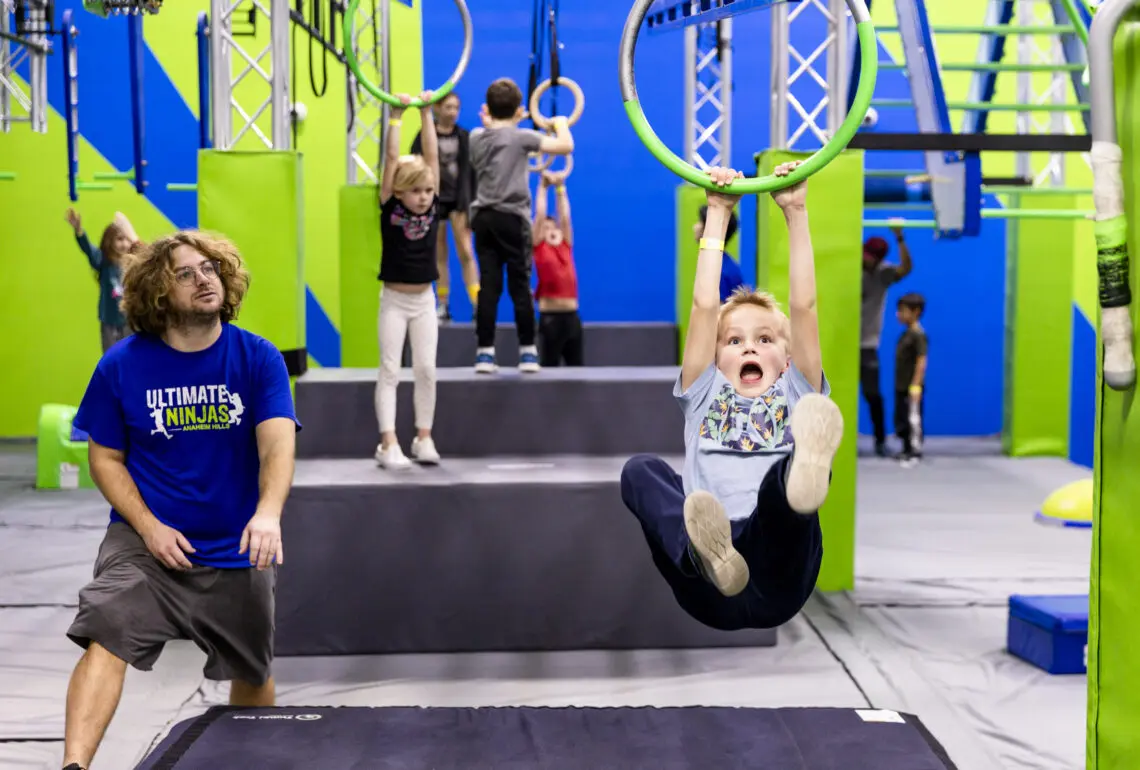 Child swinging on a green ring of a grip line, mouth open, a coach watching, and other kids on steps.